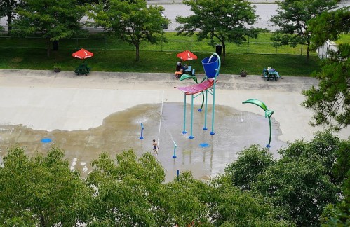 A splash pad with colorful water features, surrounded by green trees and picnic tables, on a sunny day.
