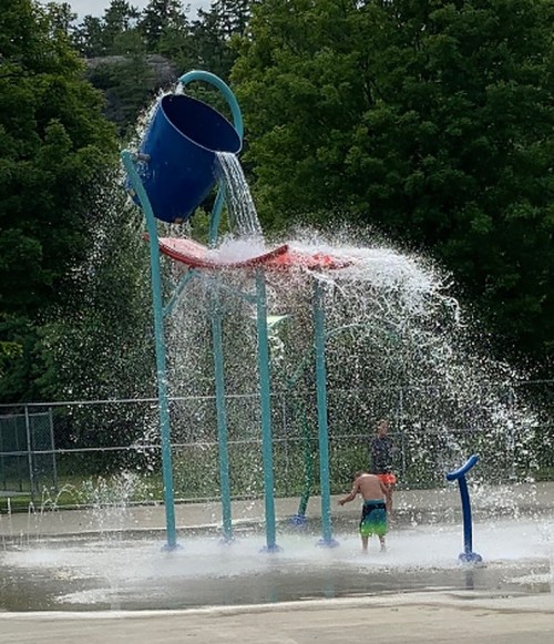 A child plays under a large water bucket pouring water at a splash pad, surrounded by greenery.