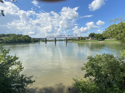 A serene river scene with a bridge, lush greenery, and fluffy clouds in a blue sky.