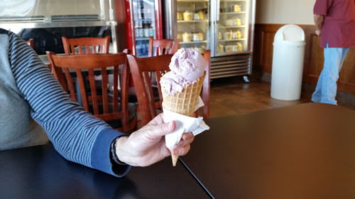 A hand holding a waffle cone with two scoops of purple ice cream, set on a black table in a café.