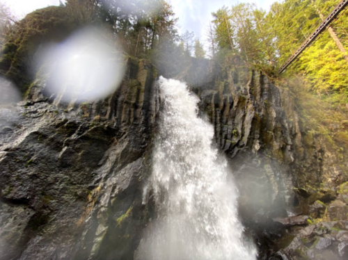 A cascading waterfall flows over rocky cliffs, surrounded by lush green trees and misty spray.