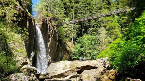 A scenic waterfall surrounded by lush greenery and rocky terrain, with a bridge overhead.