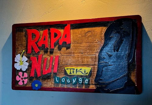 A wooden sign for "Rapa Nui Tiki Lounge" featuring bold red letters and a silhouette of a face with floral accents.
