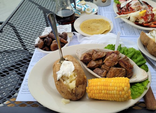 A plate with a baked potato topped with sour cream, grilled steak, corn on the cob, and a side of mushrooms.