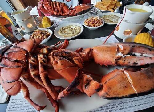 A large cooked lobster on a plate, surrounded by sides like coleslaw, corn, and sauces, with drinks in the background.