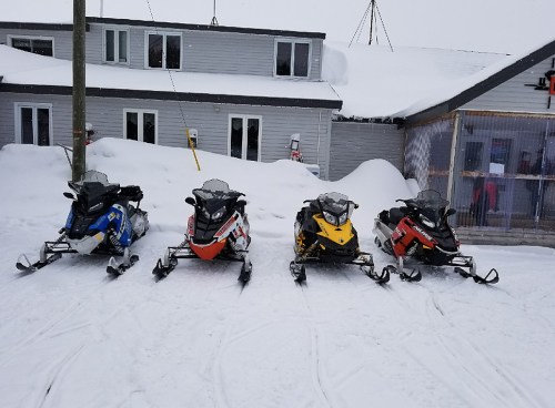 Four snowmobiles in various colors parked on a snowy surface in front of a building.