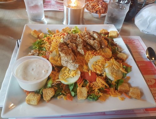 A colorful salad topped with grilled chicken, cheese, croutons, and served with a side of ranch dressing.