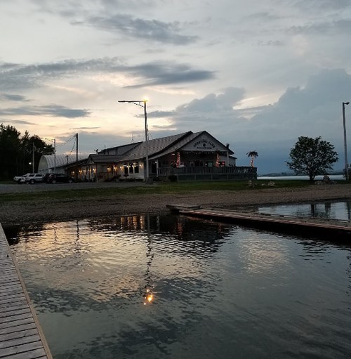 A waterfront restaurant at dusk, reflecting in calm water with a cloudy sky and a few lights glowing.