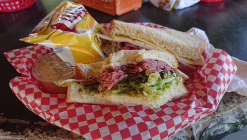 A sandwich with layers of meat and lettuce, served with a side of chips and a small container of dip, on a checkered paper.