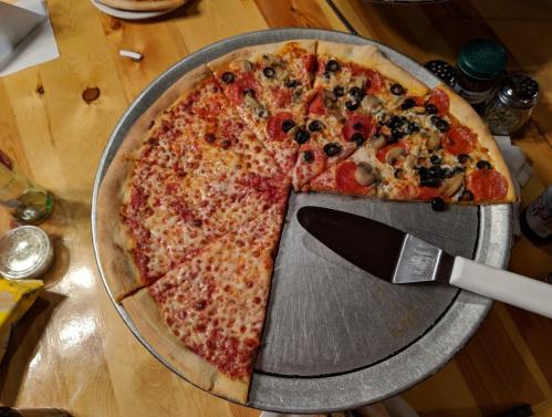 A pizza on a metal tray, half cheese and half pepperoni with olives and mushrooms, with a pizza cutter beside it.