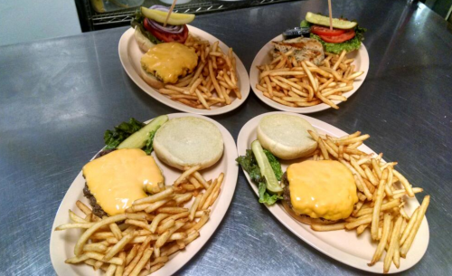 Four plates of cheeseburgers with fries, garnished with lettuce, pickles, and tomatoes, on a stainless steel surface.