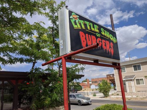 Sign for "Little Shop of Burgers" with a playful design, set against a cloudy sky and nearby buildings.