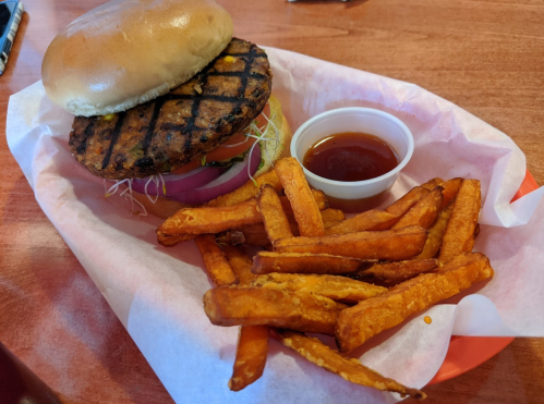 A grilled burger with lettuce and tomato, served with a side of sweet potato fries and dipping sauce on a tray.