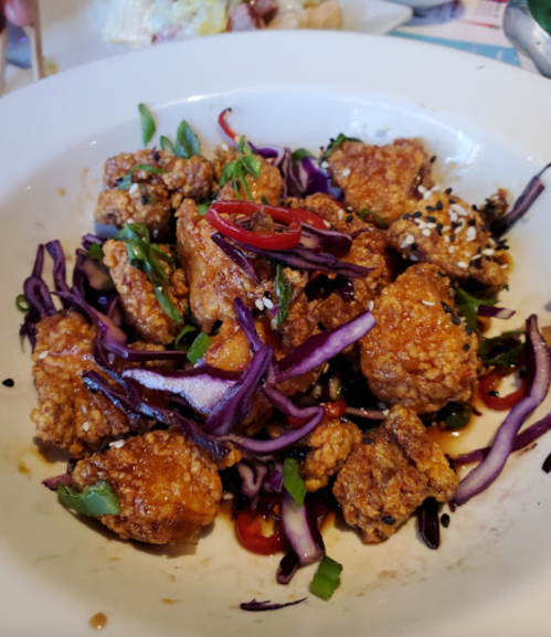 A bowl of crispy fried chicken pieces topped with purple cabbage, green onions, and red chili, drizzled with sauce.