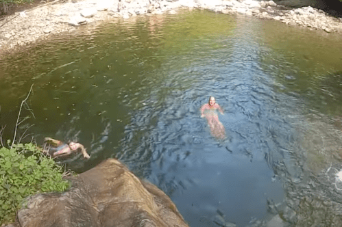 Two people swim in a natural pool surrounded by rocks and greenery, with others relaxing on the shore in the background.