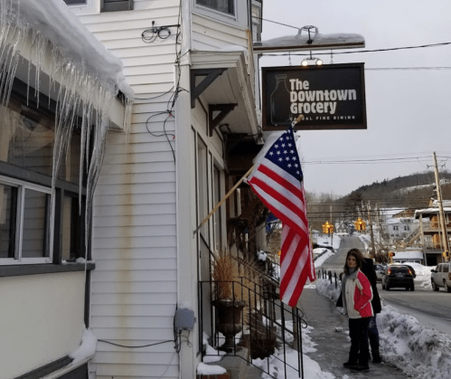A woman stands outside The Downtown Grocery, with icicles hanging and an American flag displayed. Snow covers the ground.