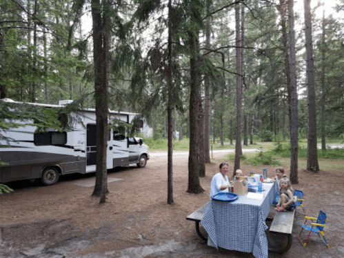 A family enjoys a meal at a picnic table in a forested campsite, with an RV parked nearby.