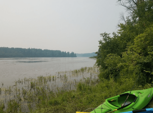 A serene river scene with a green kayak on the shore, surrounded by lush greenery and a hazy sky.