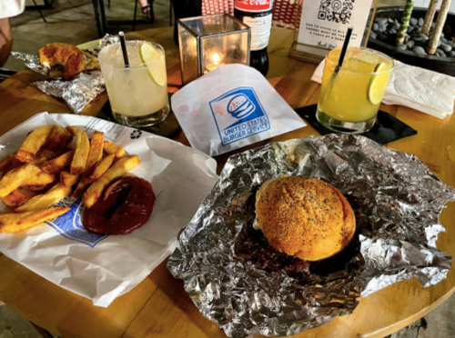 A table with a burger in foil, fries with ketchup, and two cocktails, alongside a soda and a napkin.