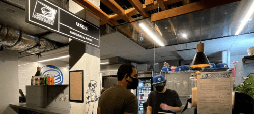 A customer in a mask interacts with a staff member at a burger counter labeled "USBS" in a casual dining setting.