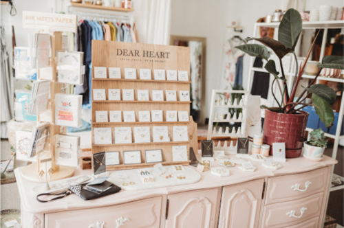 A boutique display featuring jewelry and cards on a pink dresser, with clothing and plants in the background.
