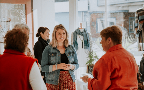 A group of women chatting in a bright store, with one smiling in a denim jacket and floral skirt.