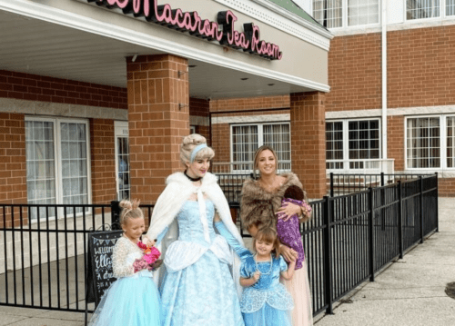 A princess in a blue gown poses with four children in costumes outside a tea room, smiling and enjoying the moment.