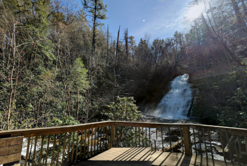 A wooden viewing platform overlooks a waterfall surrounded by lush trees and sunlight filtering through the forest.