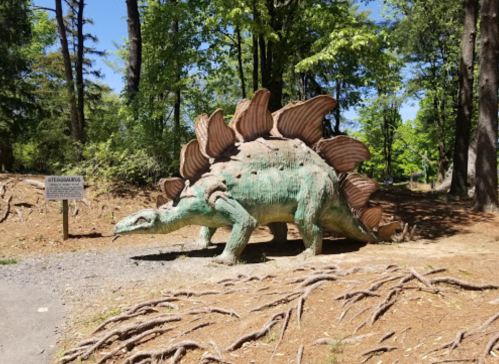 A life-sized stegosaurus sculpture in a wooded area, surrounded by trees and a sign nearby.