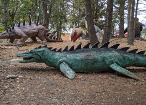 A collection of dinosaur sculptures in a wooded area, featuring various prehistoric creatures among the trees.