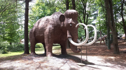 A large, brown mammoth sculpture with prominent tusks stands among trees in a park setting.