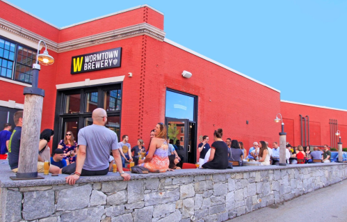 A lively outdoor scene at Wormtown Brewery, with people enjoying drinks on a sunny day by a stone wall.