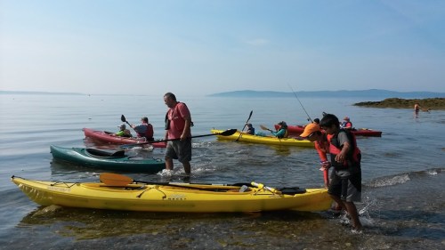 A group of people in kayaks on calm water, with some standing and others paddling near a shoreline.