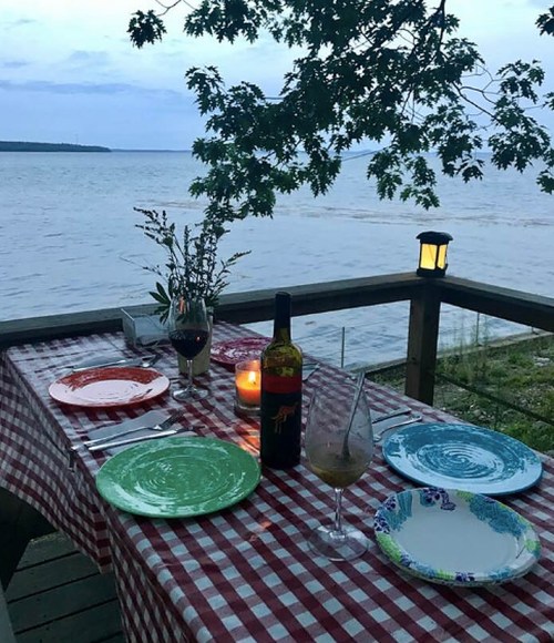 A cozy outdoor dining setup with colorful plates, wine, and a candle by the water at dusk.