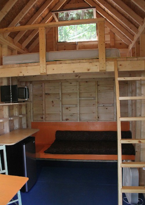 Cozy wooden cabin interior with a loft bed, seating area, kitchenette, and large window for natural light.