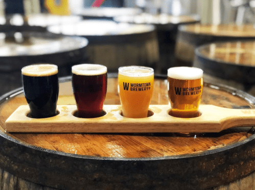 A wooden flight paddle holds four small glasses of beer on a barrel table, showcasing different beer styles.