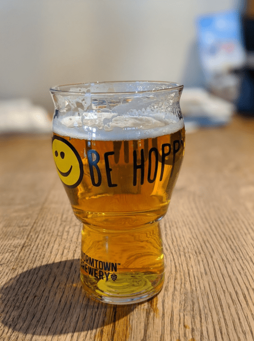 A glass of beer with a smiley face and "Be Hoppy" printed on it, sitting on a wooden table.