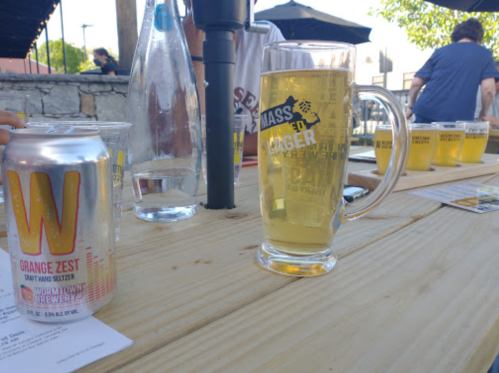 A glass of beer sits on a wooden table next to a can of orange zest hard seltzer and a flight of beers in the background.