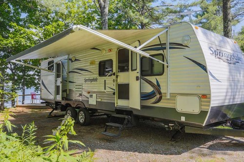 A parked travel trailer with an awning, surrounded by trees near a lake.
