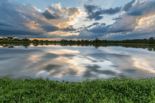 A serene lake reflects a colorful sky with clouds at sunset, surrounded by lush green grass and trees.