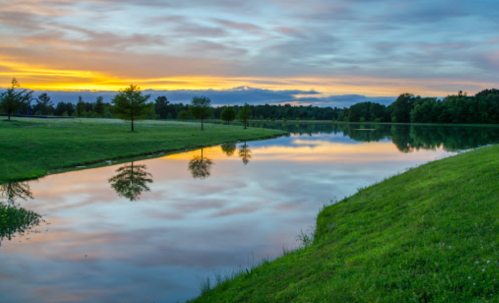 A serene lake at sunset, reflecting colorful clouds and trees along the shore, with lush green grass in the foreground.