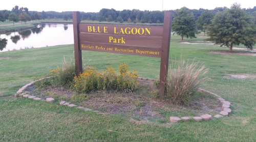 Sign for Blue Lagoon Park, featuring flowers and grass, with a pond visible in the background.