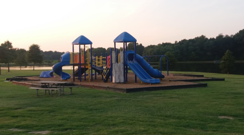 Colorful playground with slides and climbing structures, surrounded by grass and trees near a calm lake at sunset.