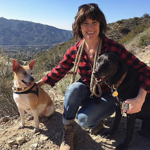 A woman in a plaid shirt kneels beside two dogs on a hiking trail with mountains in the background.