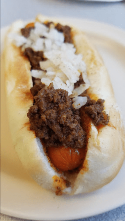 A hot dog in a bun topped with chili and chopped onions on a light-colored plate.
