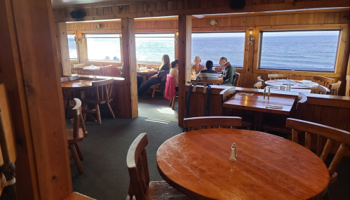 Cozy wooden restaurant interior with ocean views, featuring tables and diners enjoying their meals.