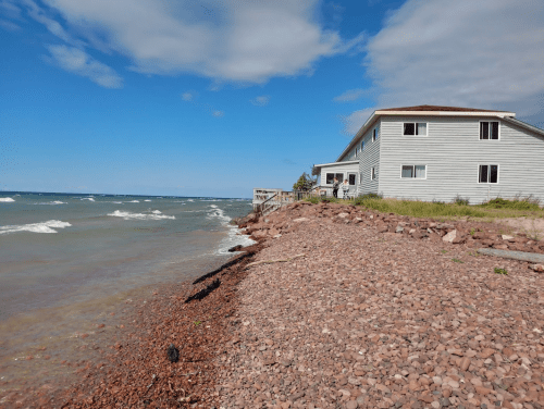 A house by the shore with rocky beach and waves, under a partly cloudy sky.