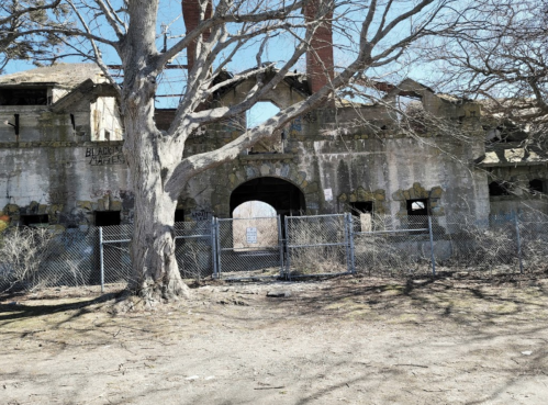 Abandoned building with crumbling walls, a large tree in front, and a chain-link fence surrounding the area.