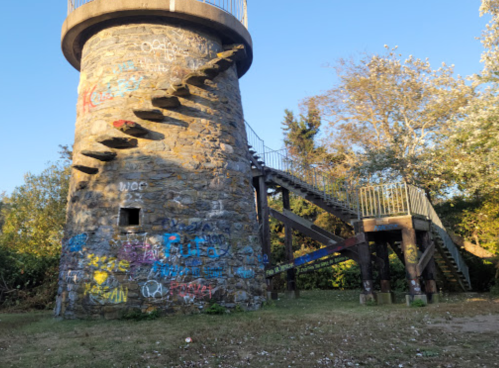 A stone tower with a spiral staircase, covered in colorful graffiti, surrounded by trees and a clear blue sky.
