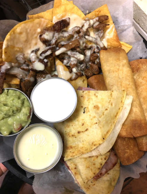 A platter of nachos with cheese, meat, and sauces, alongside taquitos, guacamole, and creamy dips.
